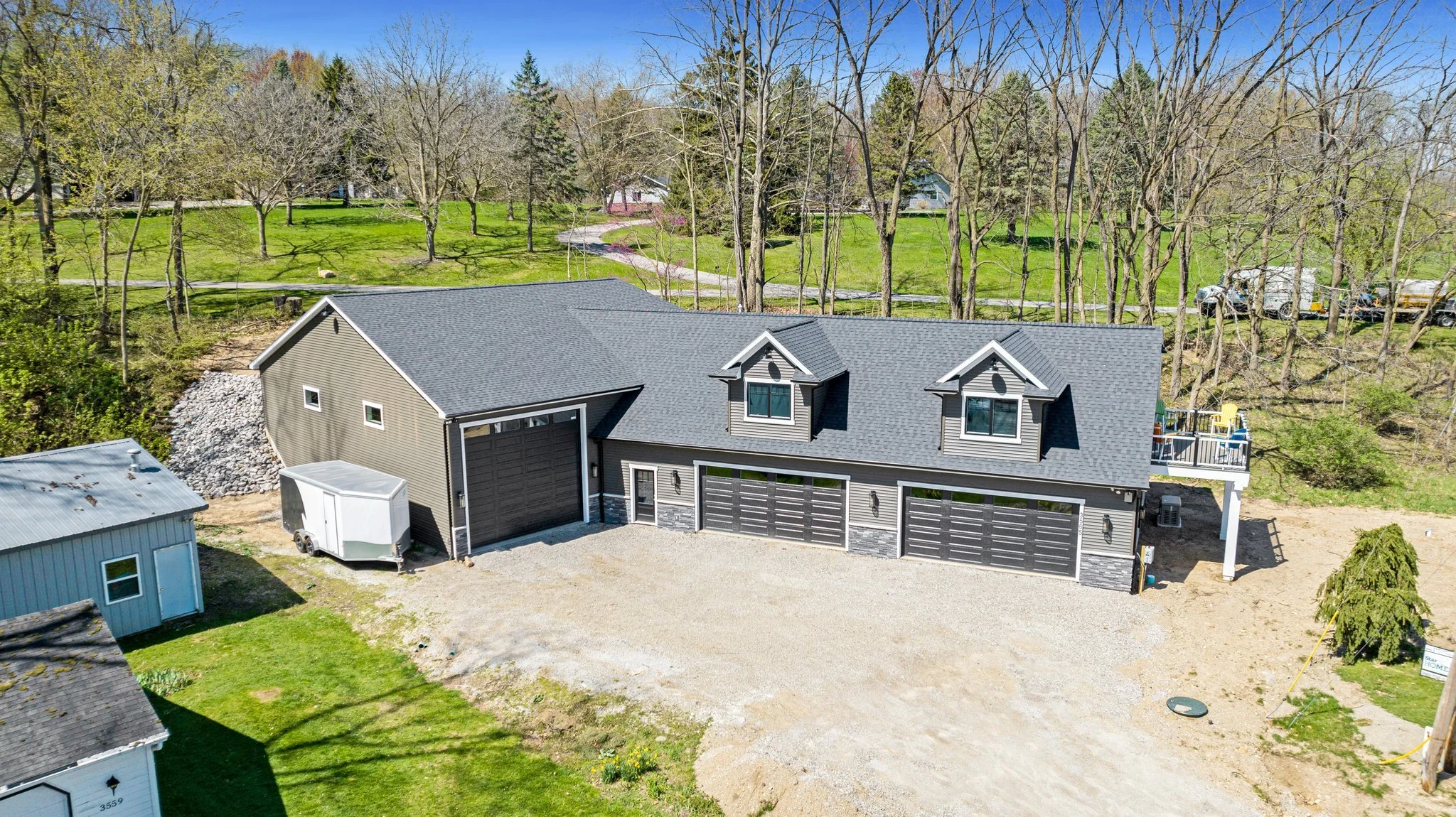 A newly built gray garage with multiple garage doors, dormer windows, a gravel driveway, and a small storage shed nearby, surrounded by trees and greenery.