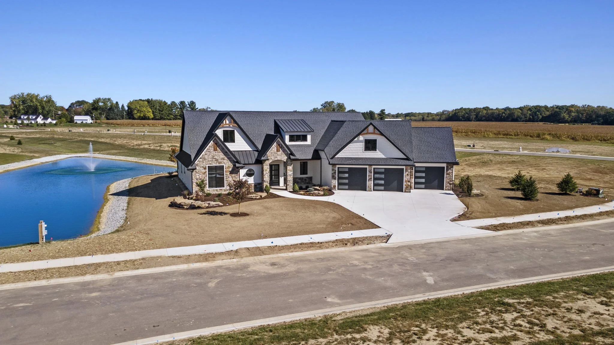 A new two-story house with a stone and white exterior, black roof, and three garage doors, situated next to a pond with a fountain, and landscaped with small trees and shrubs.