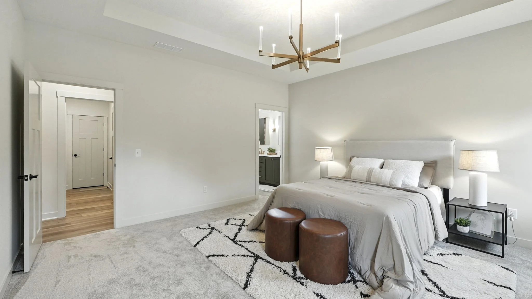 A bedroom with a neutral-colored bed, two bedside lamps, two brown leather ottomans, a black metal side table with a small plant, white walls, a modern gold chandelier, and an open doorway leading to a bathroom