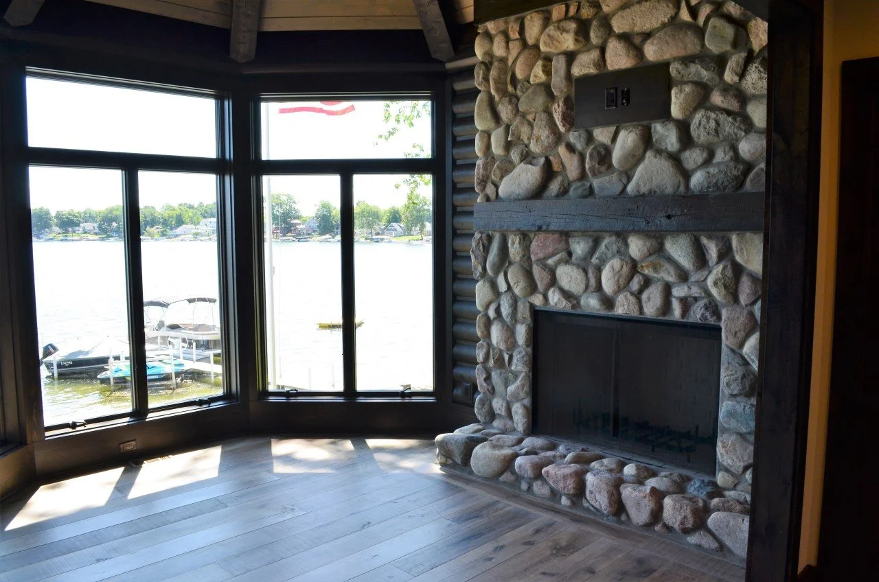 Living room with large windows overlooking a lake, with boats docked outside, and a stone fireplace inside.