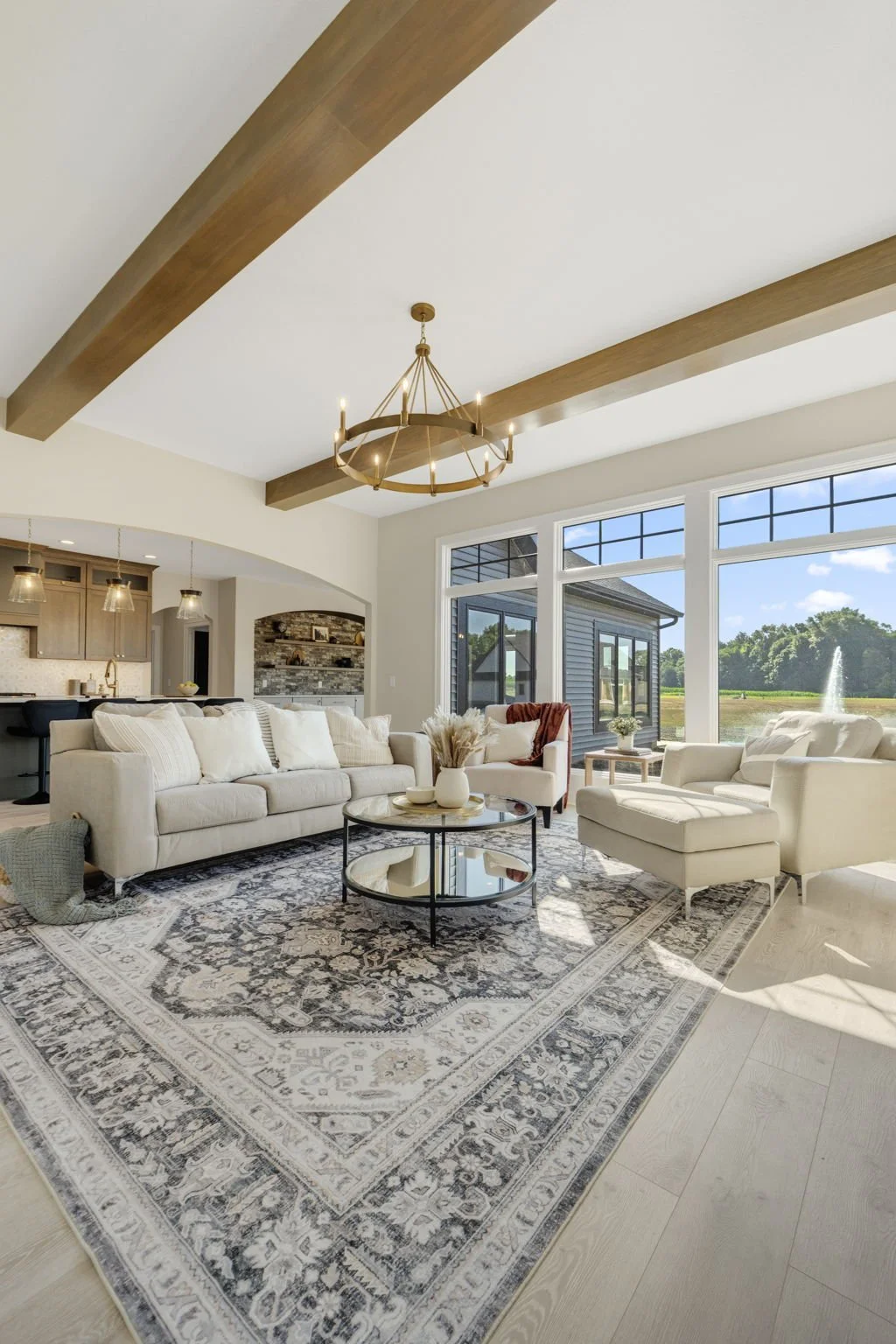Bright living room with large windows, cream-colored sofa and armchair, round glass coffee table, patterned area rug, and wooden ceiling beams with a modern chandelier.