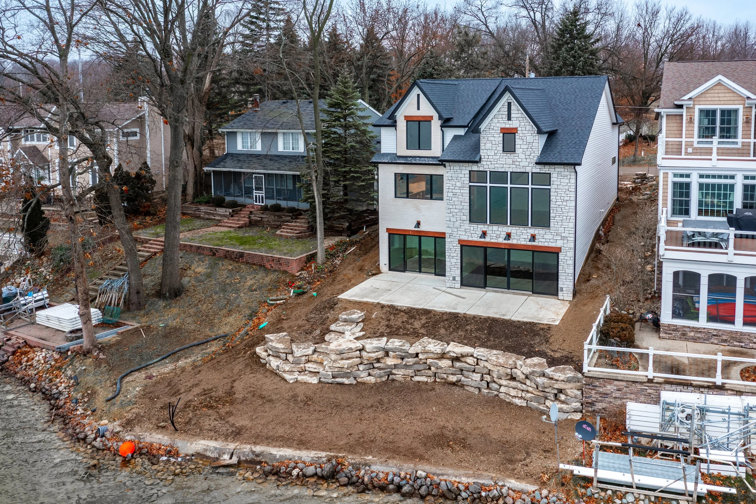 Newly built modern multi-story house with large windows and a stone facade, adjacent to neighboring houses, with a rocky hillside and a partially landscaped yard under construction.