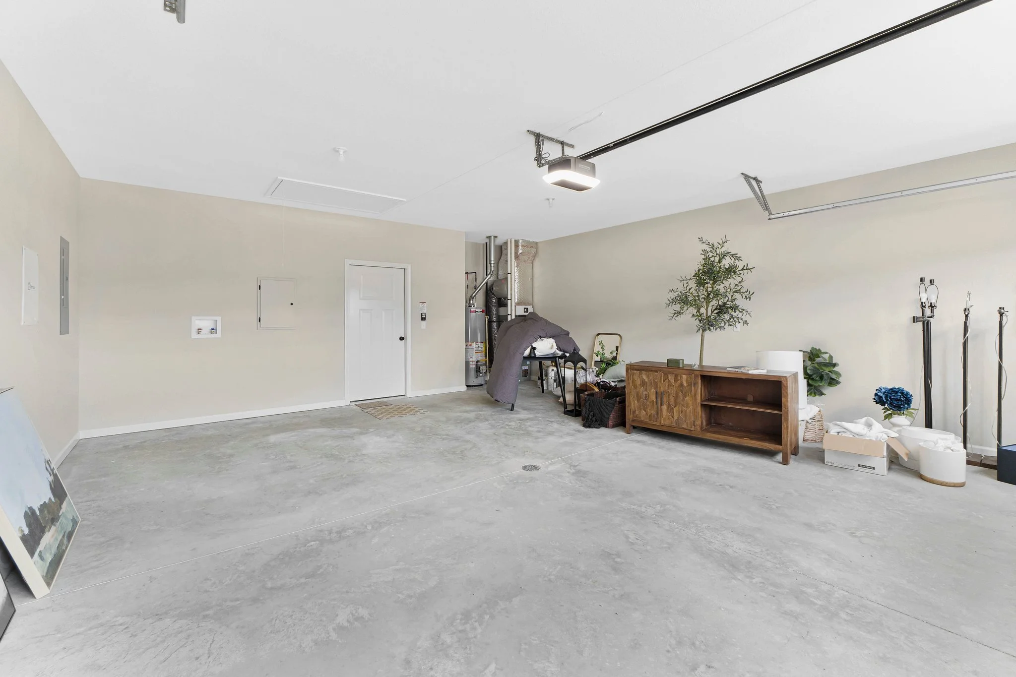 Empty garage with beige walls, concrete floor, and some furniture, plants, and boxes near the right wall. There is a garage door opener mounted on the ceiling.