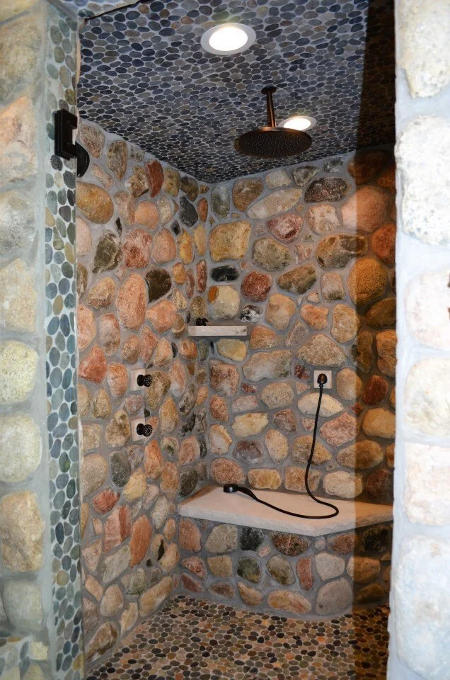 A rustic stone shower with pebble-tile floor and ceiling, featuring a rainfall showerhead, built-in corner bench, and multiple plumbing fixtures.