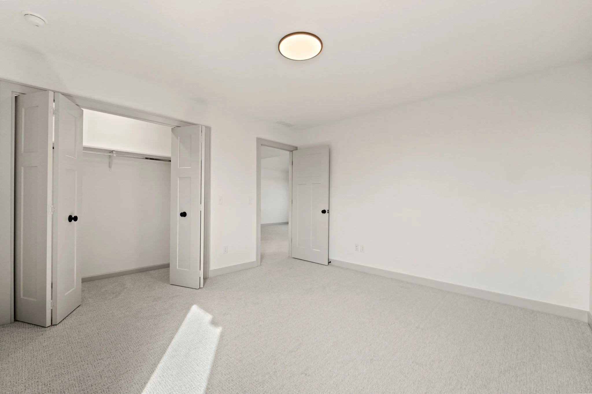 A clean, empty bedroom with white walls, beige carpet, open closet doors, and a ceiling light.