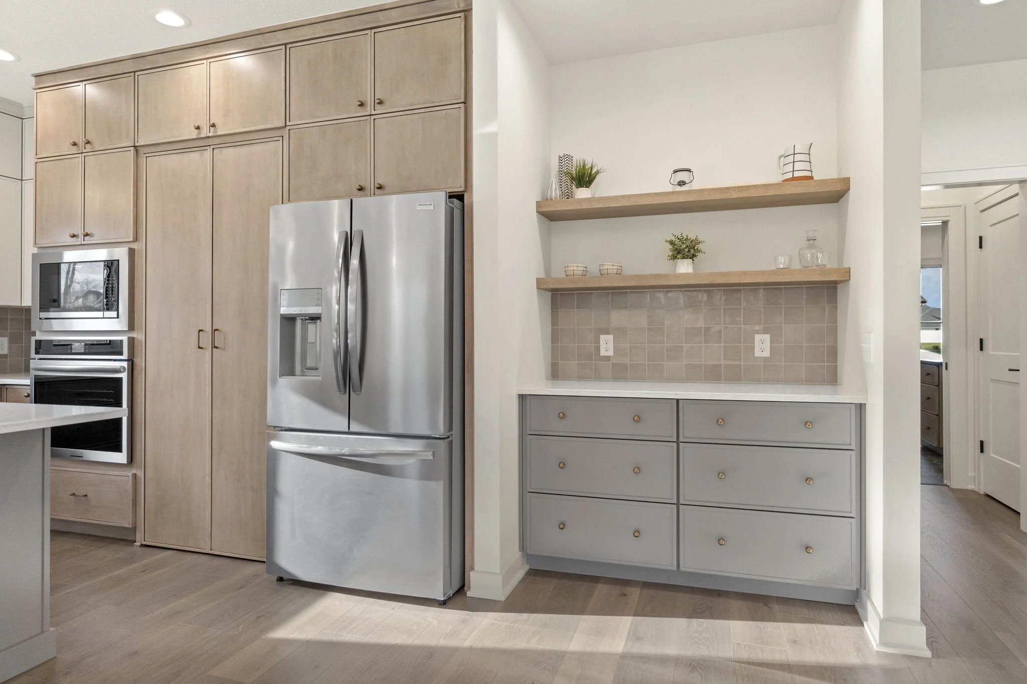 Modern kitchen with light wood cabinets, stainless steel refrigerator, gray drawers, and open wooden shelves with decorative items.