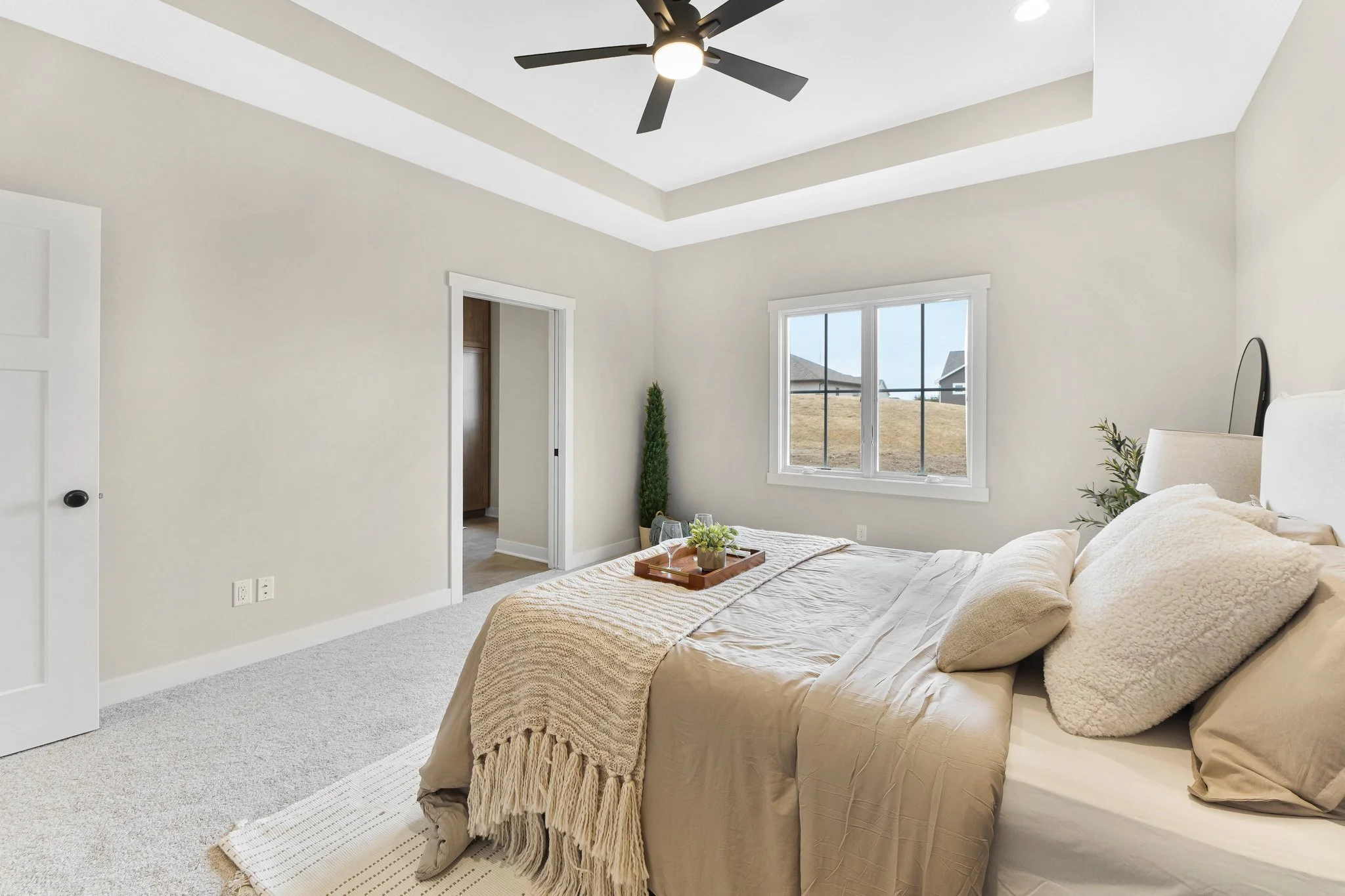 A bedroom with a large bed covered in beige bedding and pillows, a tray with plants on the bed, a window showing an outdoor view, a ceiling fan, and light-colored walls and carpet.
