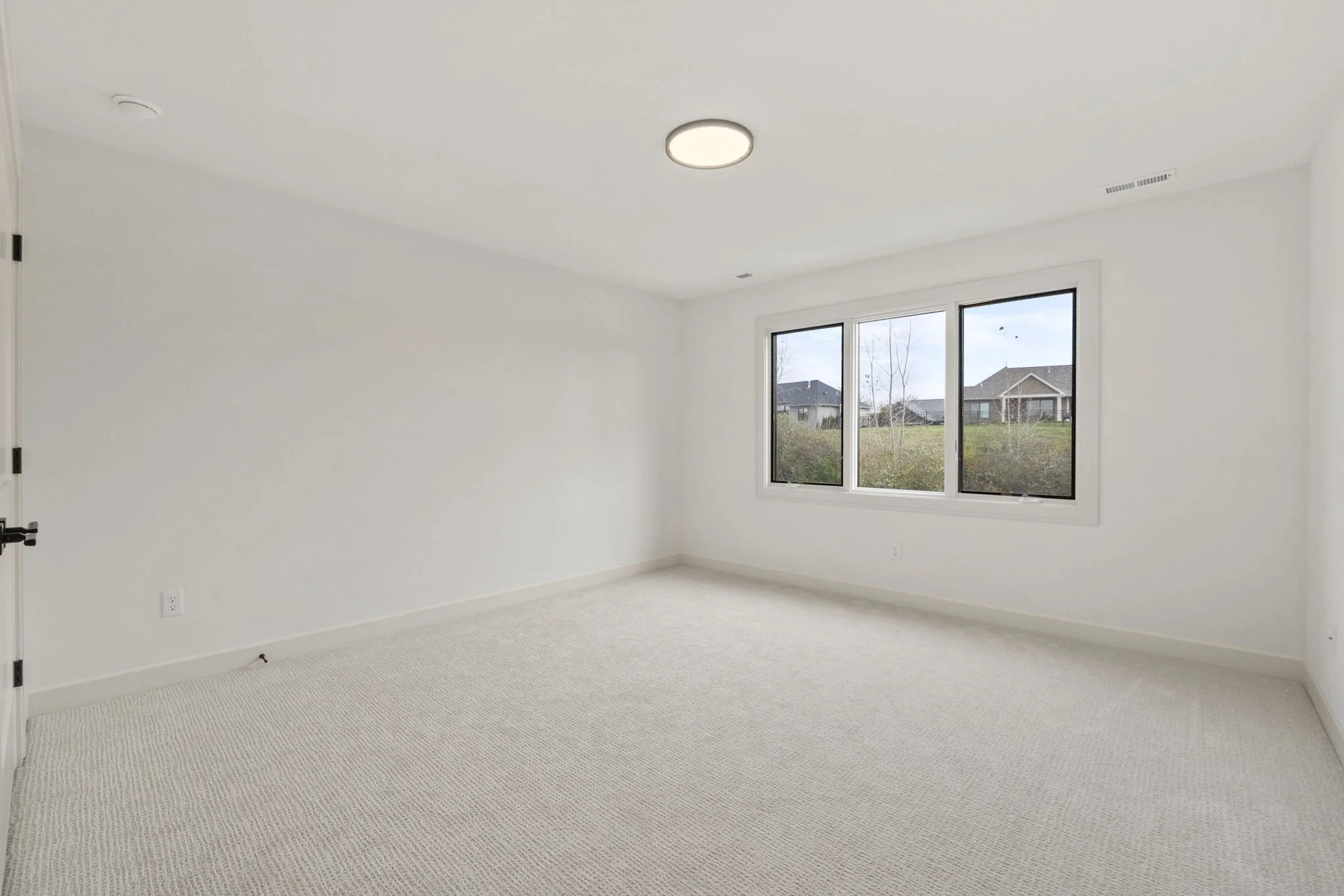 Empty white room with large window showing houses outside, beige carpet, ceiling light, and white walls.
