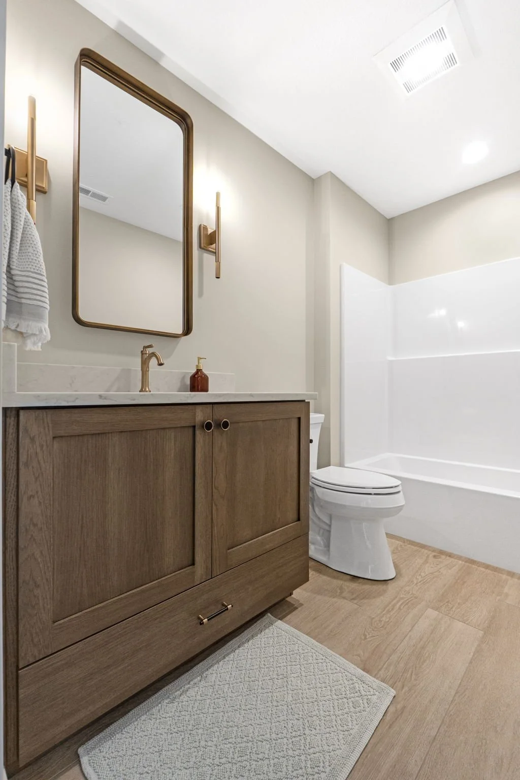 Bathroom with wooden vanity, white countertop, mirror with wooden frame, wall lights, beige walls, white toilet, and bathtub with white surround.