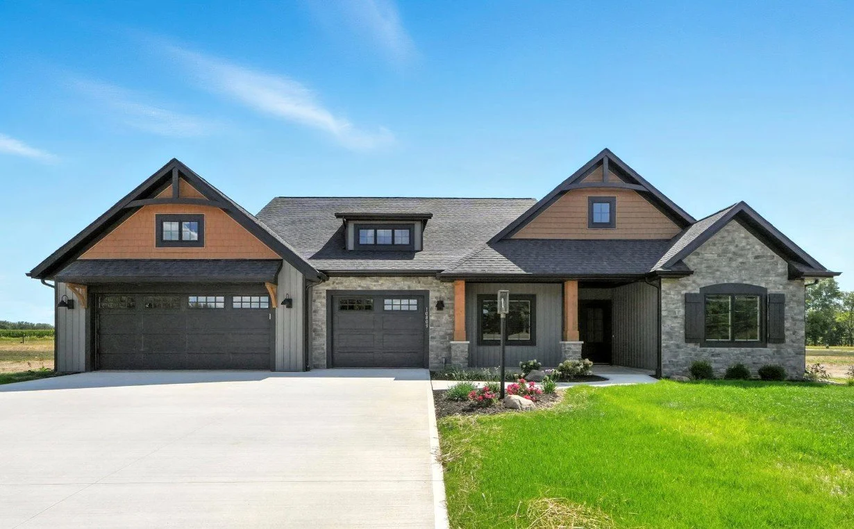 Modern single-family house with stone and wood siding, gray garage doors, a small front porch, and a well-maintained front yard with green grass and flowers.