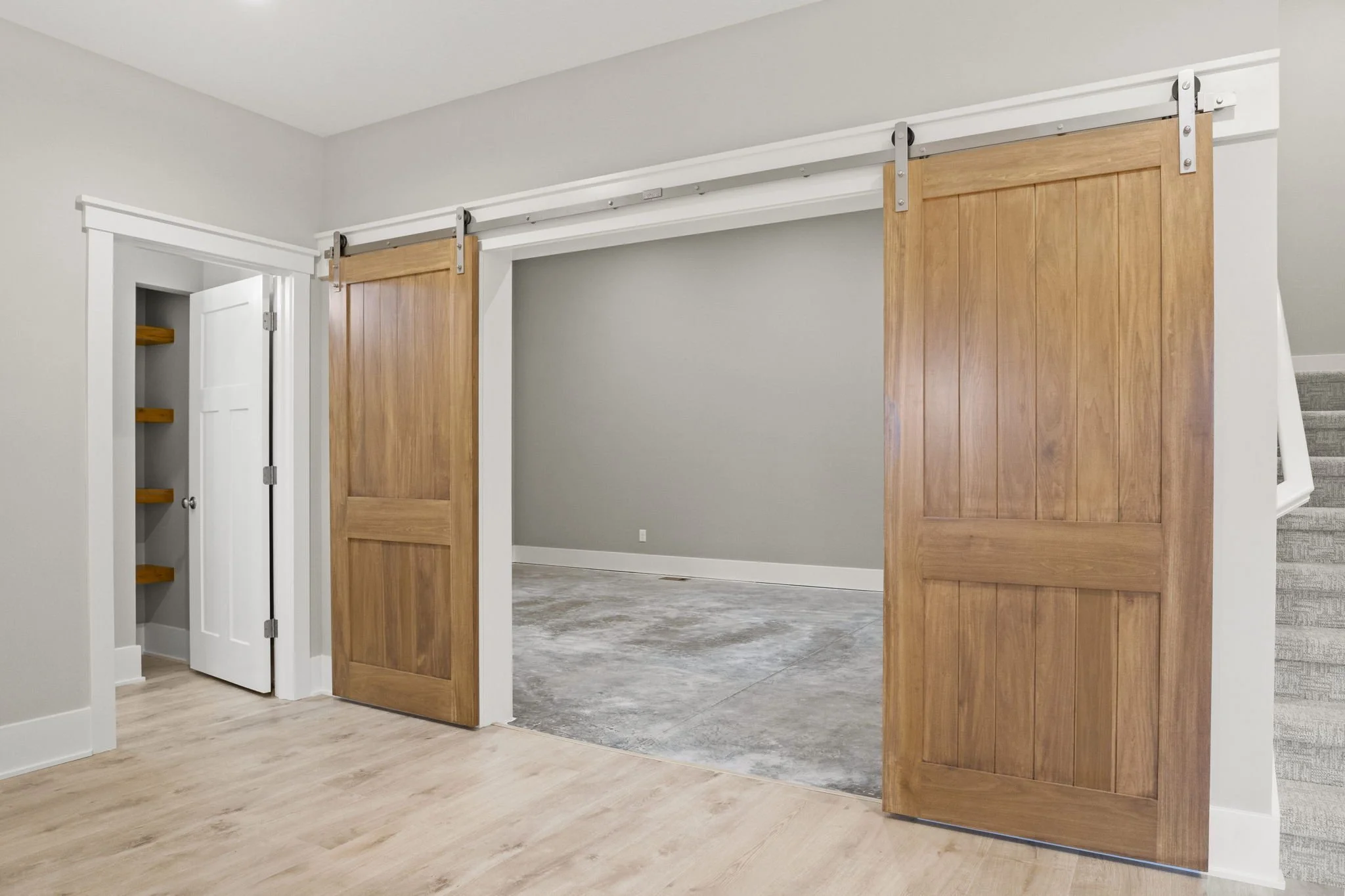 Interior view of a room with a set of sliding barn doors made of wood, partially open, revealing a storage closet with shelves and a white door, and a staircase on the right side of the image.