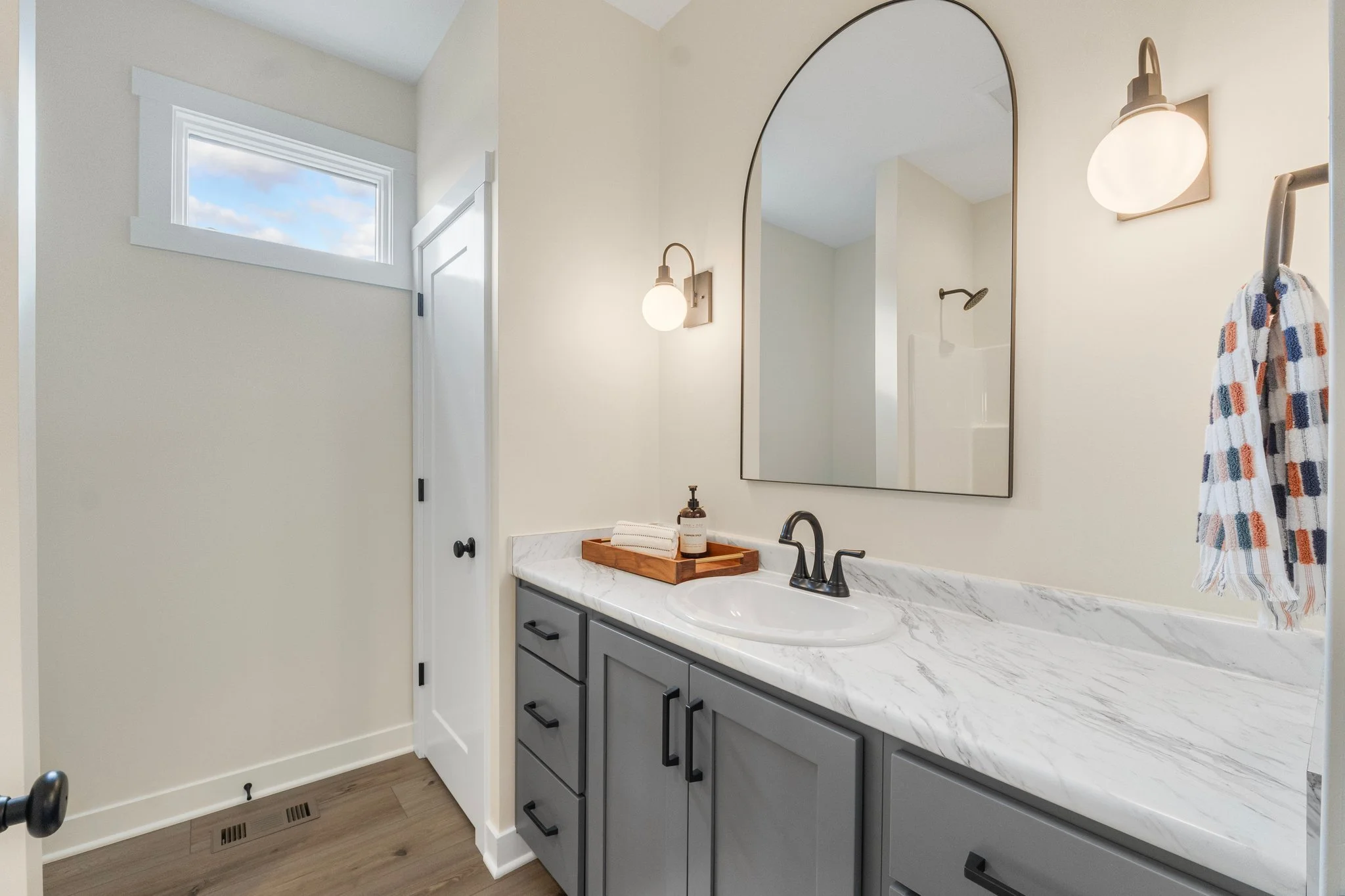 Modern bathroom with a marble countertop, gray cabinets, a large mirror, a wall-mounted light, a small window, a towel ring with a checkered towel, and a shower in the background.