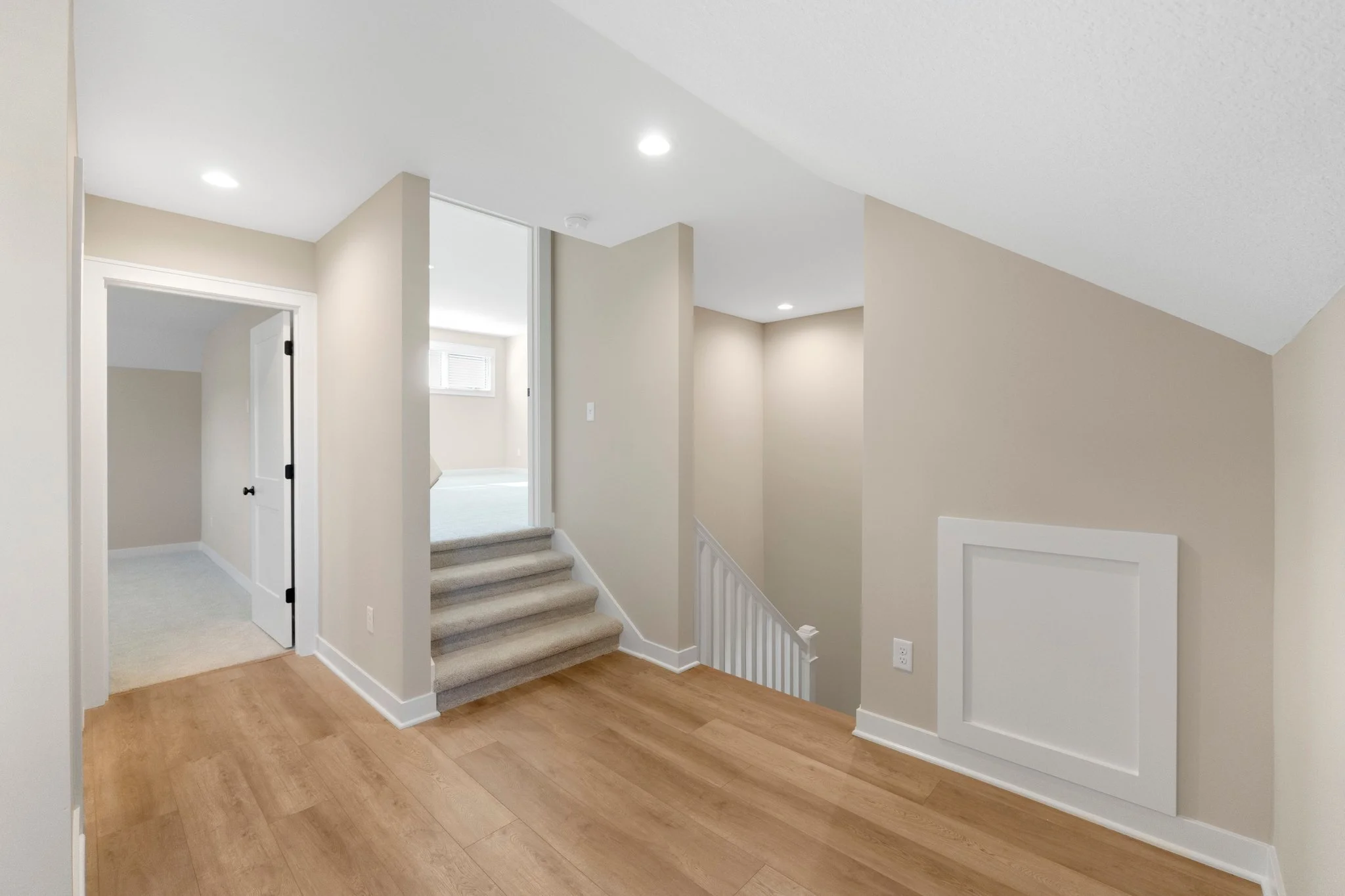 Interior of a home's upper floor showing beige walls, wood flooring, carpeted stairs, and multiple rooms with white trim and trim panel on the wall.