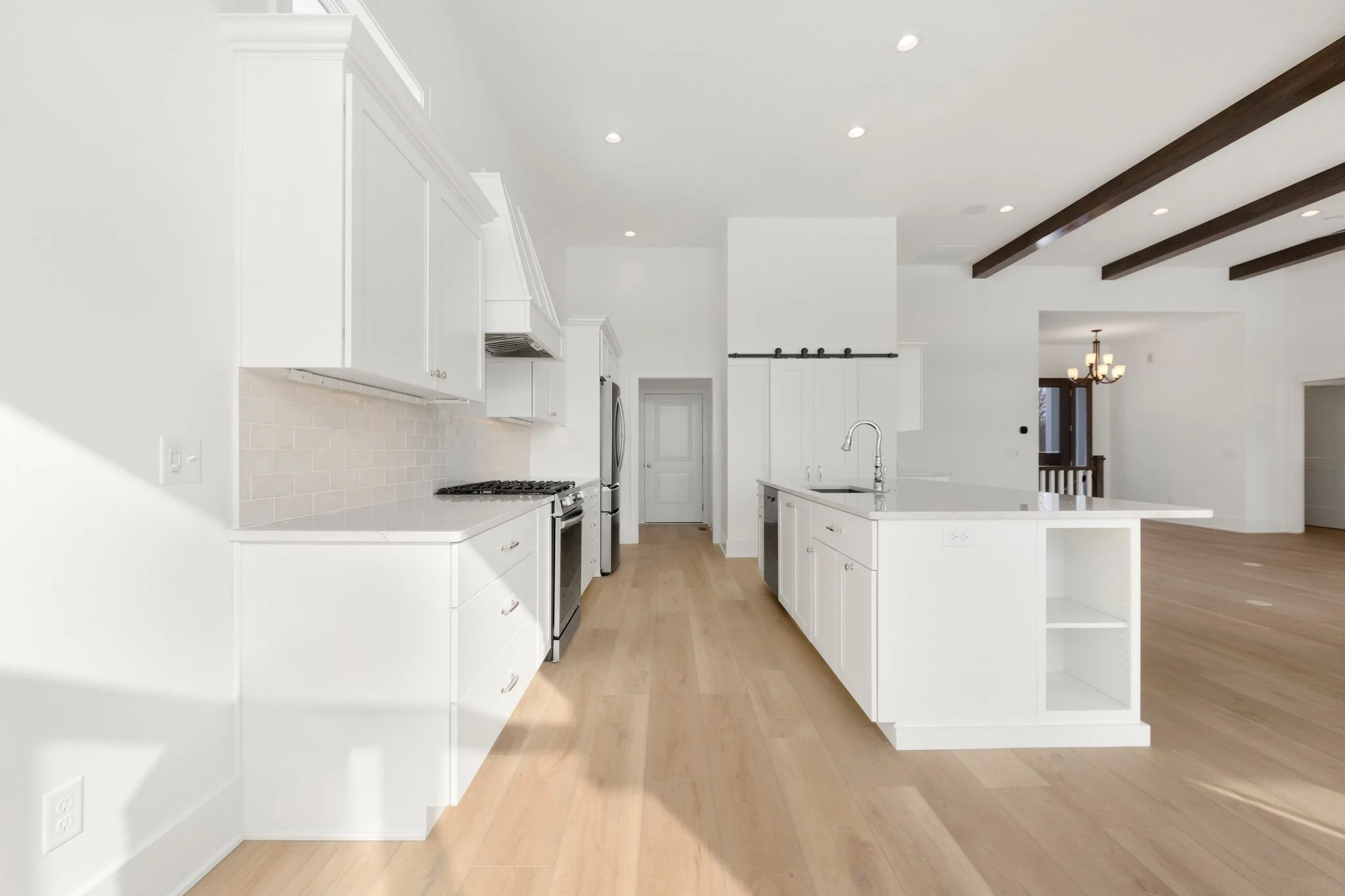 Modern white kitchen with island, stainless steel appliances, light wood flooring, and exposed dark wood ceiling beams.