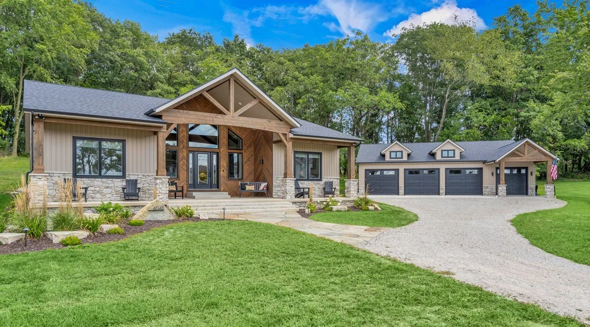 Large modern house with stone and wood exterior, surrounded by green lawn and trees, with a gravel driveway leading to a four-car garage.