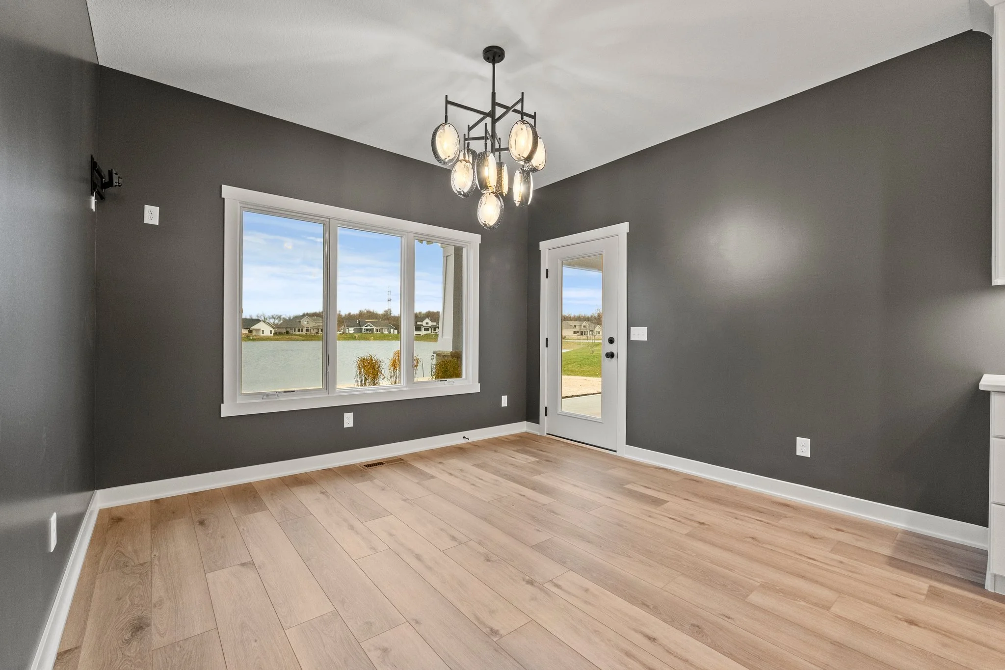Empty room with gray walls, hardwood floor, large window showing outdoor view, door with glass panel, and modern ceiling light fixture.