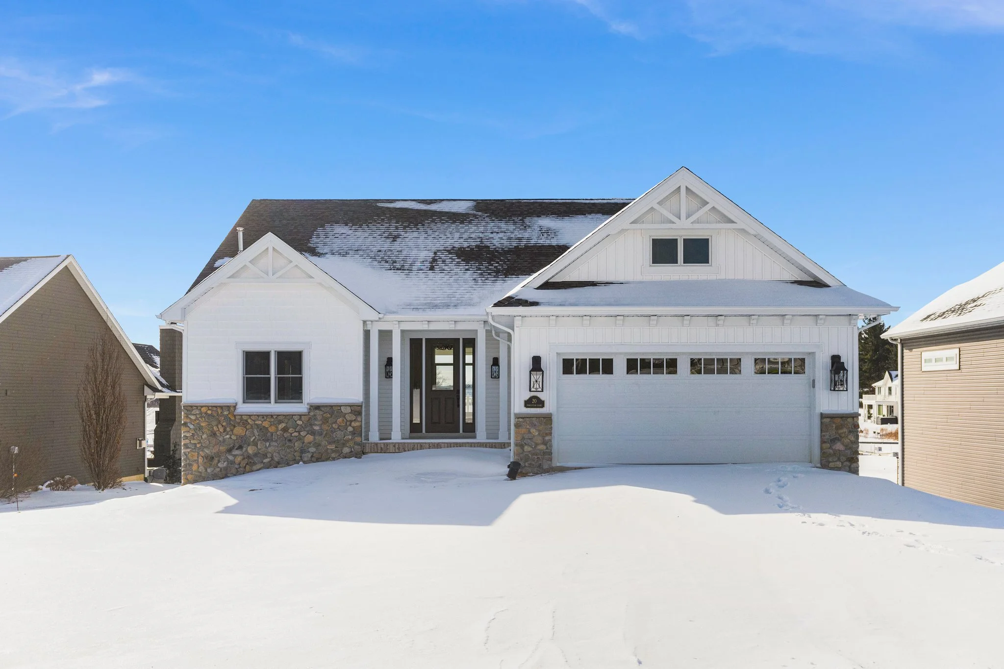 White house with stone accents and a two-car garage, snow on the ground and roof, clear blue sky.