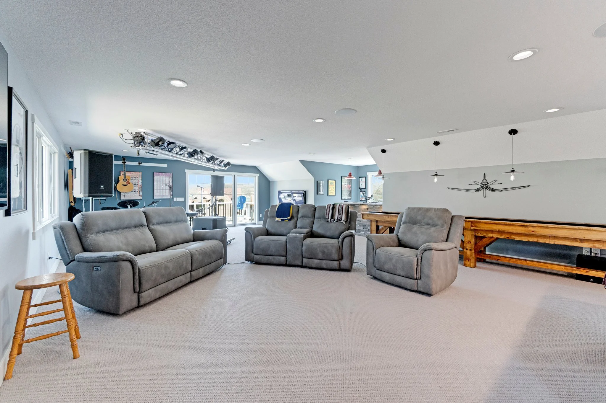 Spacious living room with three gray recliner sofas, wooden stools, and large windows opening to a deck. Music equipment, guitars, and a TV are visible in the background.