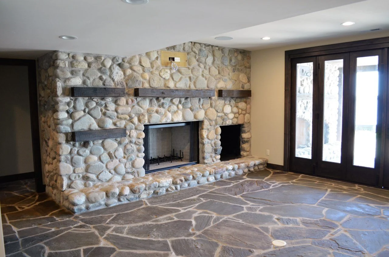 A living room with a stone fireplace, dark wood shelves above it, and a stone and wood floor. There is a set of closed glass doors on the right.