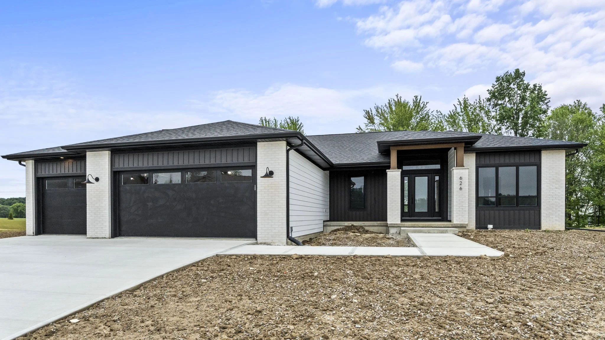 New modern house with black doors and windows, white brick and black siding exterior, concrete driveway, and front yard with dirt and some grass, under a partly cloudy sky.