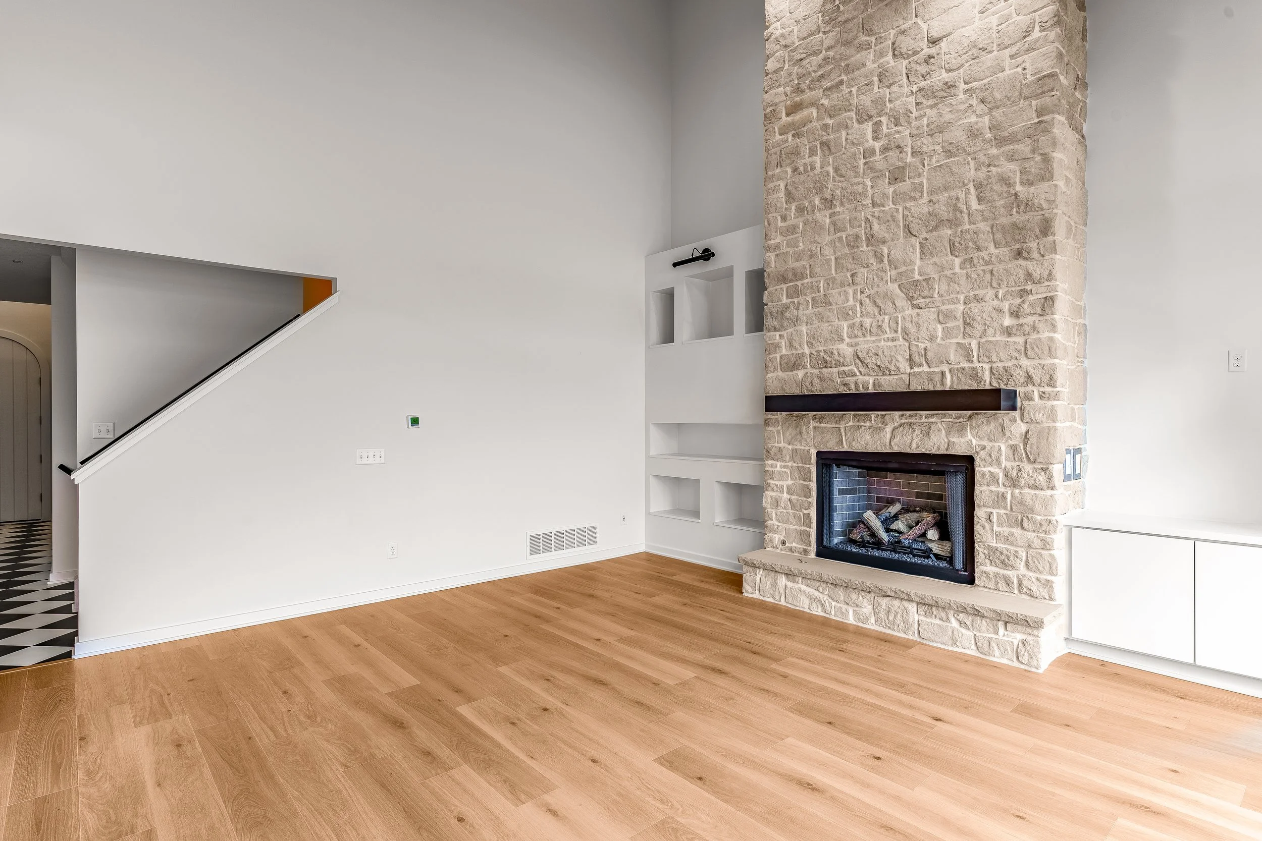 Empty living room with light wood floors, a stone fireplace, built-in white shelves, and a staircase with a black handrail.
