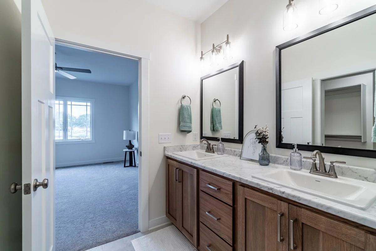 Master bathroom with double sink vanity, marble countertop, large mirror, and wall-mounted light fixtures, adjacent to a carpeted bedroom with window and ceiling fan.