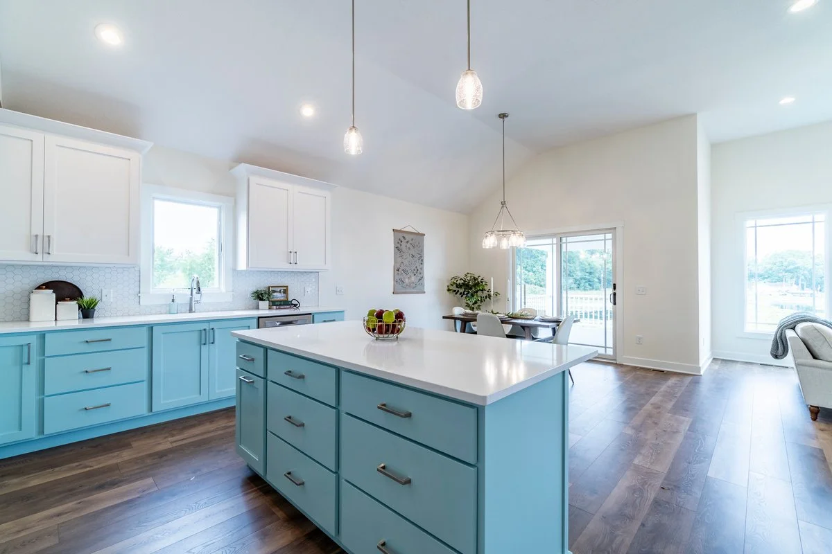 Bright modern kitchen with white upper cabinets, light blue lower cabinets, a central island, and hardwood floors. There are pendant lights hanging from the ceiling, a window above the sink, and a sliding glass door leading to an outdoor area.