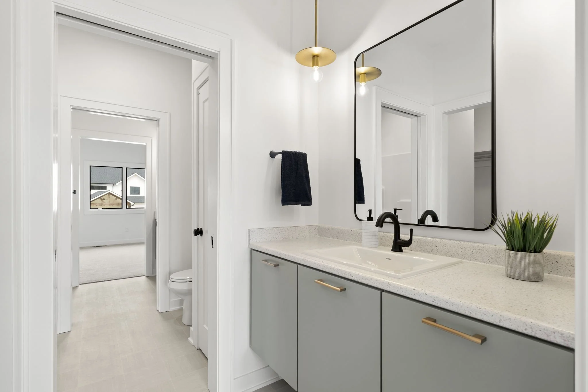 Modern bathroom with a light-colored speckled countertop, black fixtures, and a large mirror. There is a potted plant on the counter and a black towel on the wall. Visible through an open door is a room with a window showing outdoor houses and a bed.