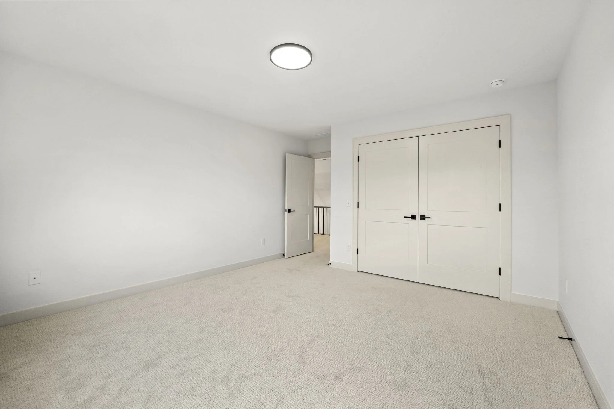 Empty room with white walls, beige carpet, a double-door closet, and an open door leading to a hallway.