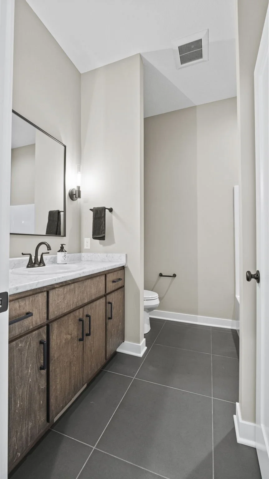 Modern bathroom with a wooden vanity, marble countertop, black fixtures, gray floor tiles, a mirror, a towel, and a nearby toilet.