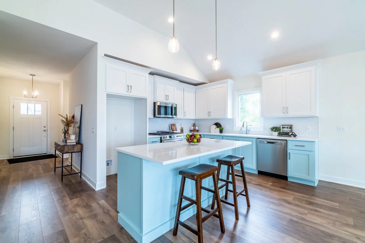 A bright, modern kitchen with white cabinets, a light blue kitchen island, wooden stools, stainless steel appliances, and hardwood flooring.