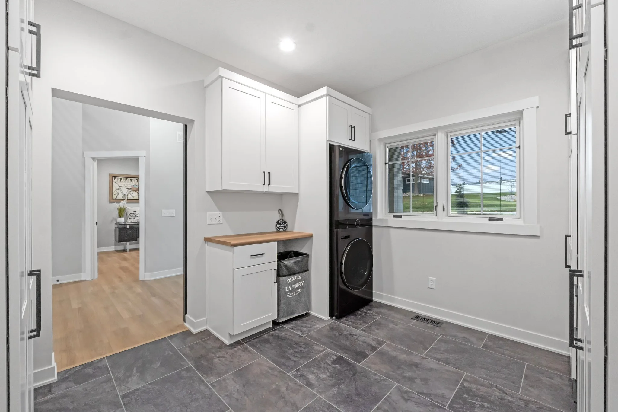 Laundry room with black stacked washer and dryer, white cabinets, gray tile floor, window showing outdoor scene, and laundry hamper labeled 'Deluxe Laundry Service'.