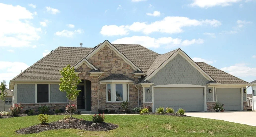 A modern suburban house with stone and siding exterior, a gabled roof, and three-car garage, surrounded by a well-maintained lawn and landscaping.