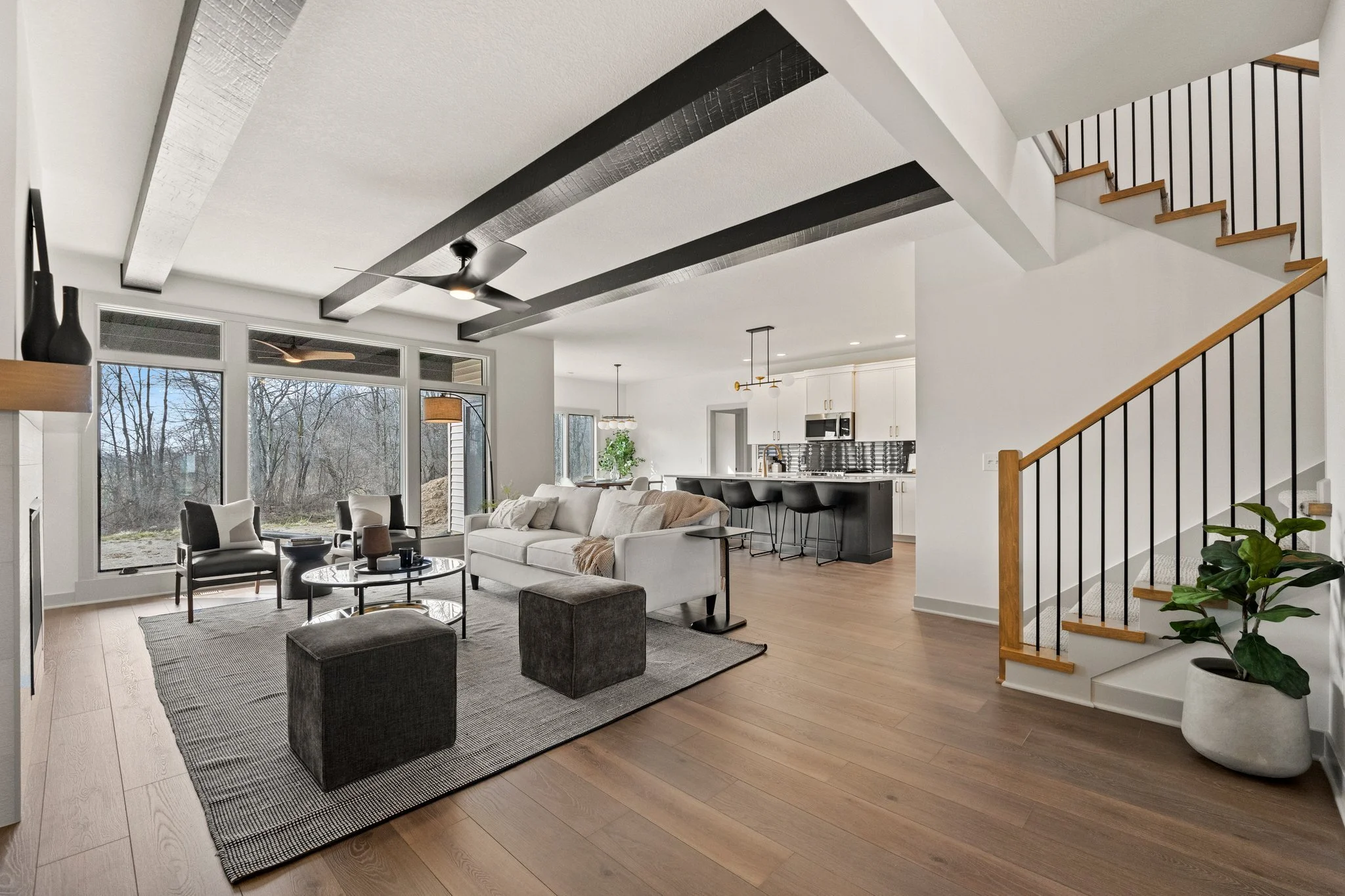 Modern open-concept living room with large windows, white walls, a white sofa, black armchairs, a gray rug, and a staircase with wooden handrail and black balusters.