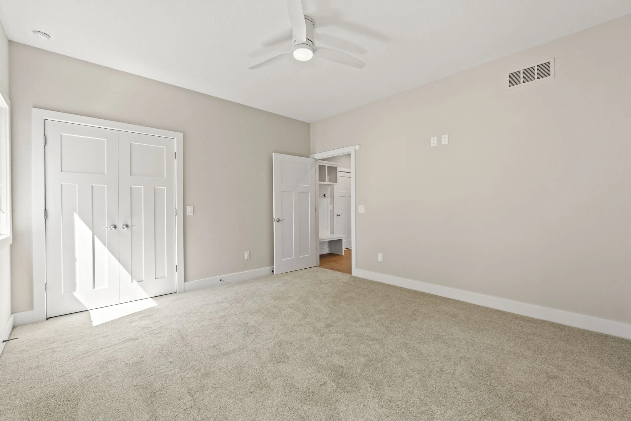 Empty beige carpeted room with white walls, double closet doors, a ceiling fan, and a doorway leading to another room with a small built-in storage bench.