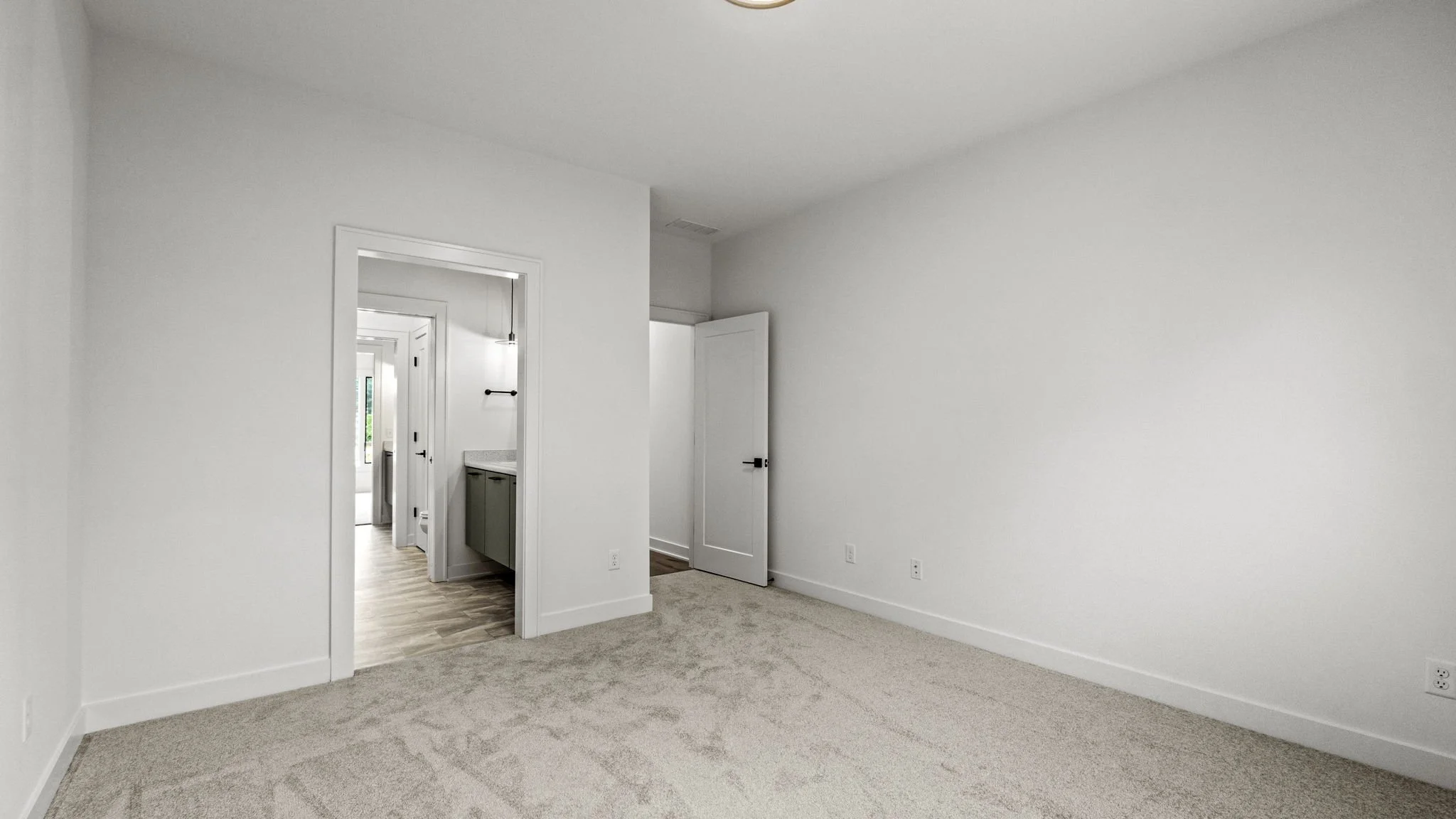Empty room with beige carpet, white walls, and white trim. There are three doors, one open leading to a hallway with wood flooring and a bathroom with green cabinets. The room has multiple electrical outlets.