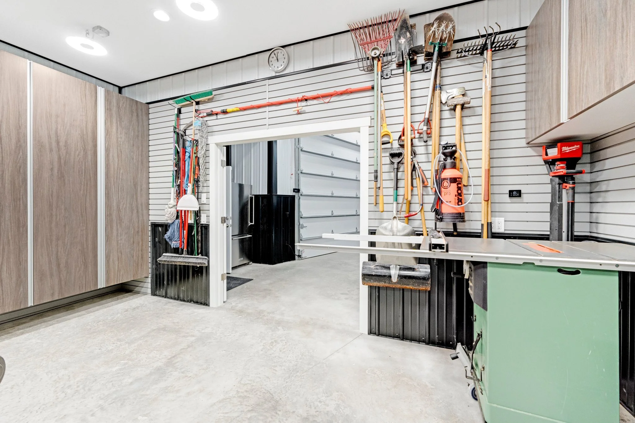 Well-organized garage workshop with tools hanging on a slatwall, including shovels, rakes, and a weed trimmer. A workbench with a green cabinet, a saw, and a roll-up garage door in the background.