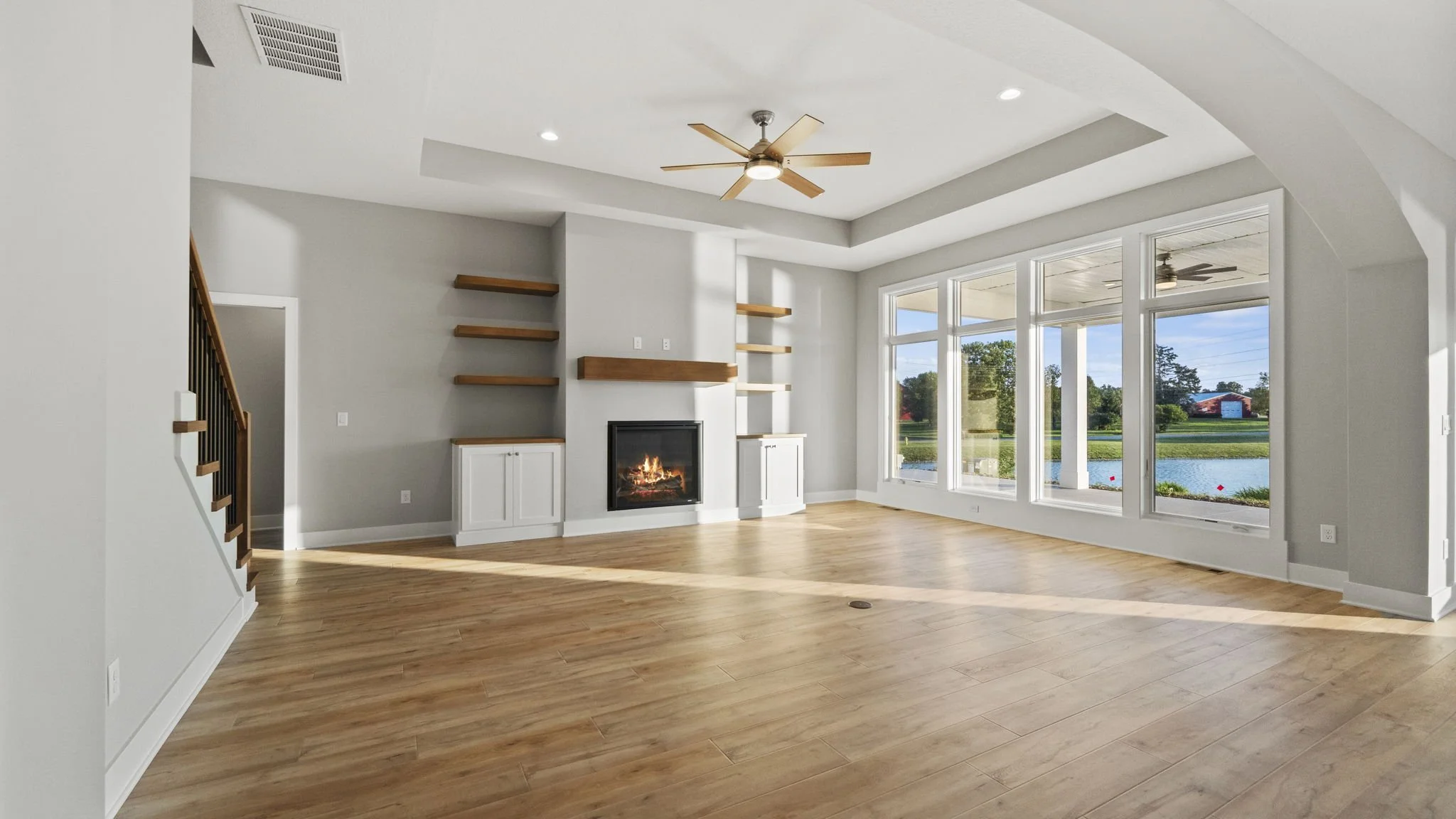 Empty living room with large windows overlooking a pond, hardwood floors, a fireplace, a ceiling fan, and built-in shelves.