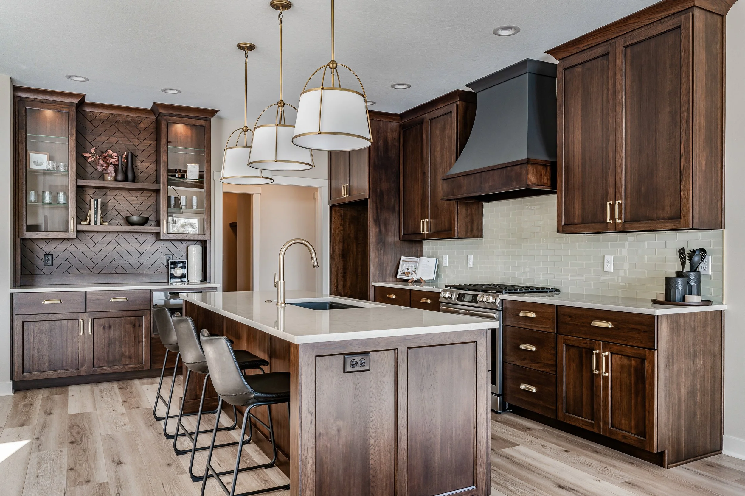 Modern kitchen with dark wood cabinets, white island with seating, gray range hood, beige tile backsplash, and pendant lights.