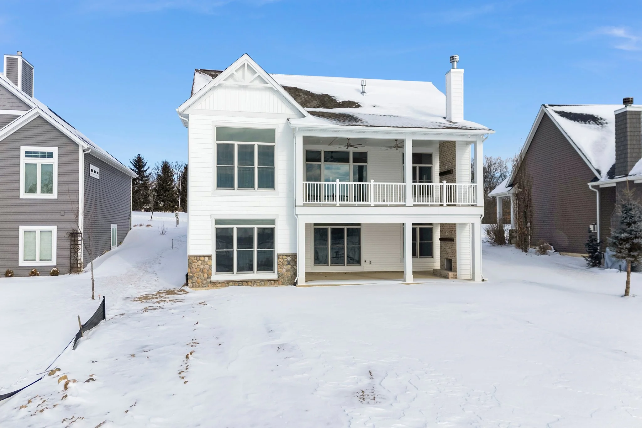 A two-story white house with a stone foundation, large windows, a balcony, and snow-covered yard, flanked by neighboring houses on a winter day.