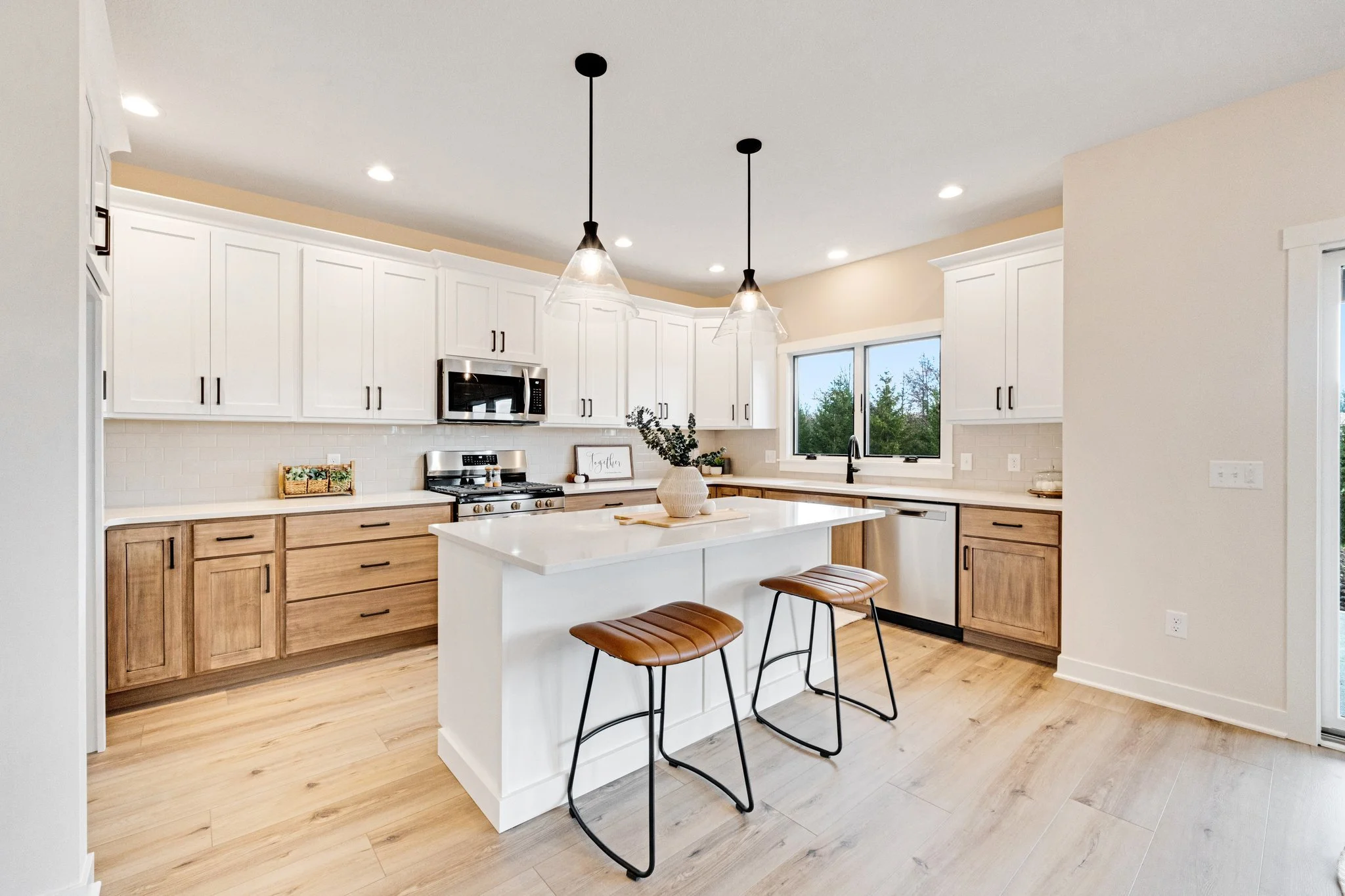 Modern kitchen with white cabinets, wooden lower cabinets, a central island with a white countertop, pendant lights, and a window view of trees outside.