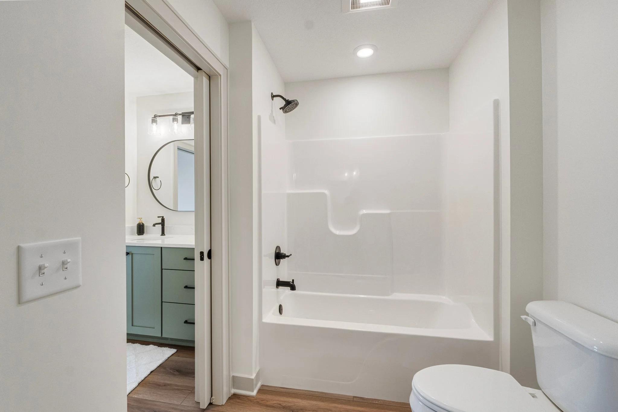Bathroom with white walls, a shower/tub combination with a dark showerhead, a white toilet, and a view into a vanity area with a mirror and a light fixture.