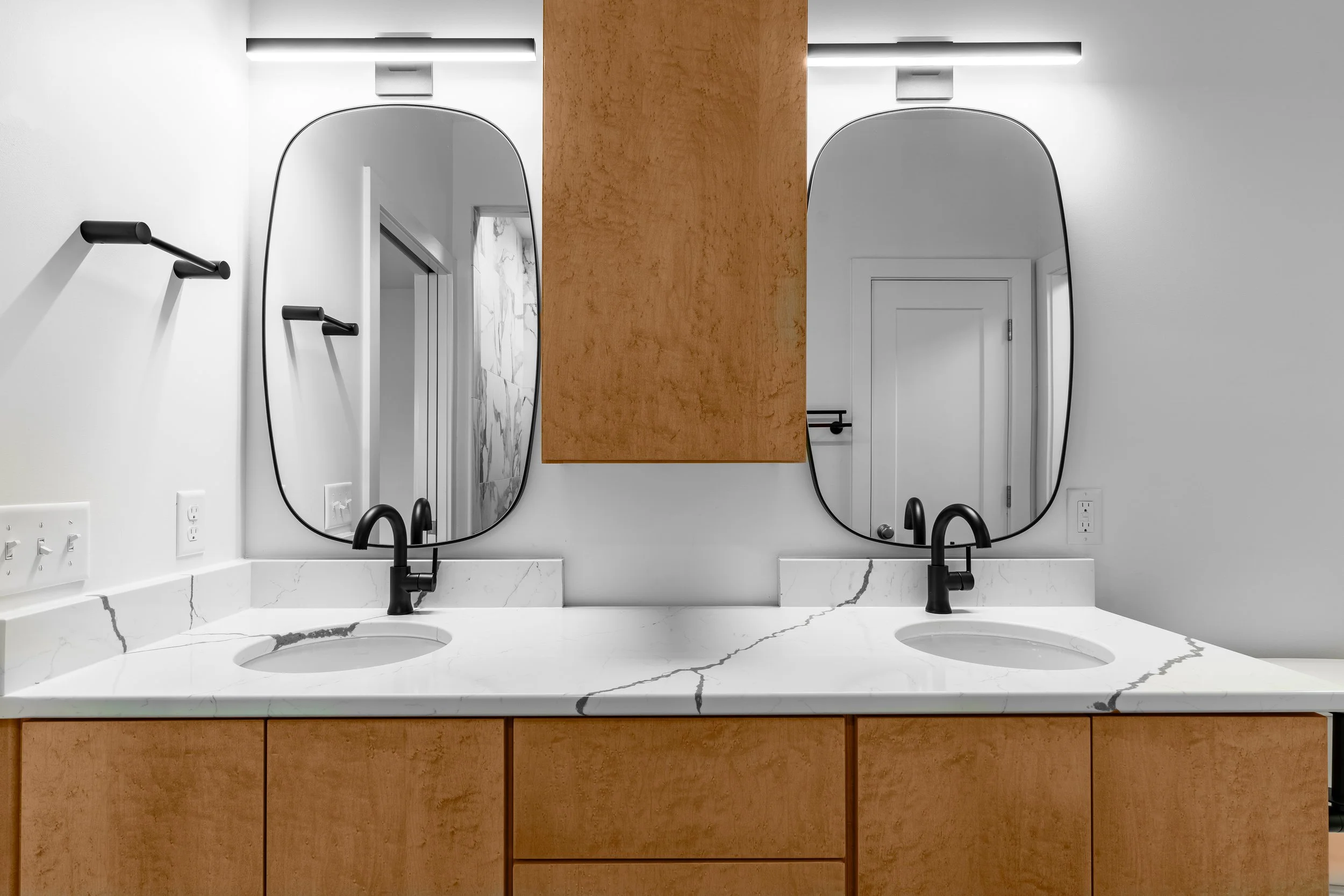 Bathroom double vanity with black faucets, white marble countertop, wooden cabinets, two large oval mirrors, and a wood panel hanging between the mirrors.