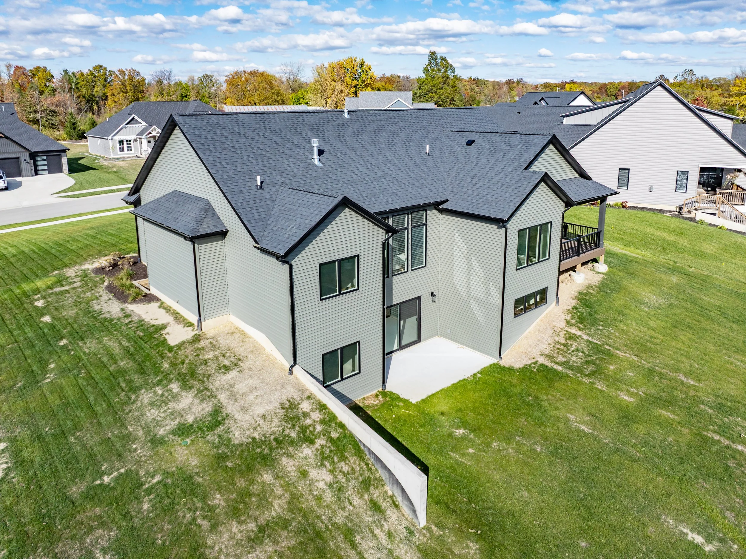 Aerial view of a modern two-story house with gray siding, black roof, and small backyard patio, located in a suburban neighborhood with similar houses and green lawns.