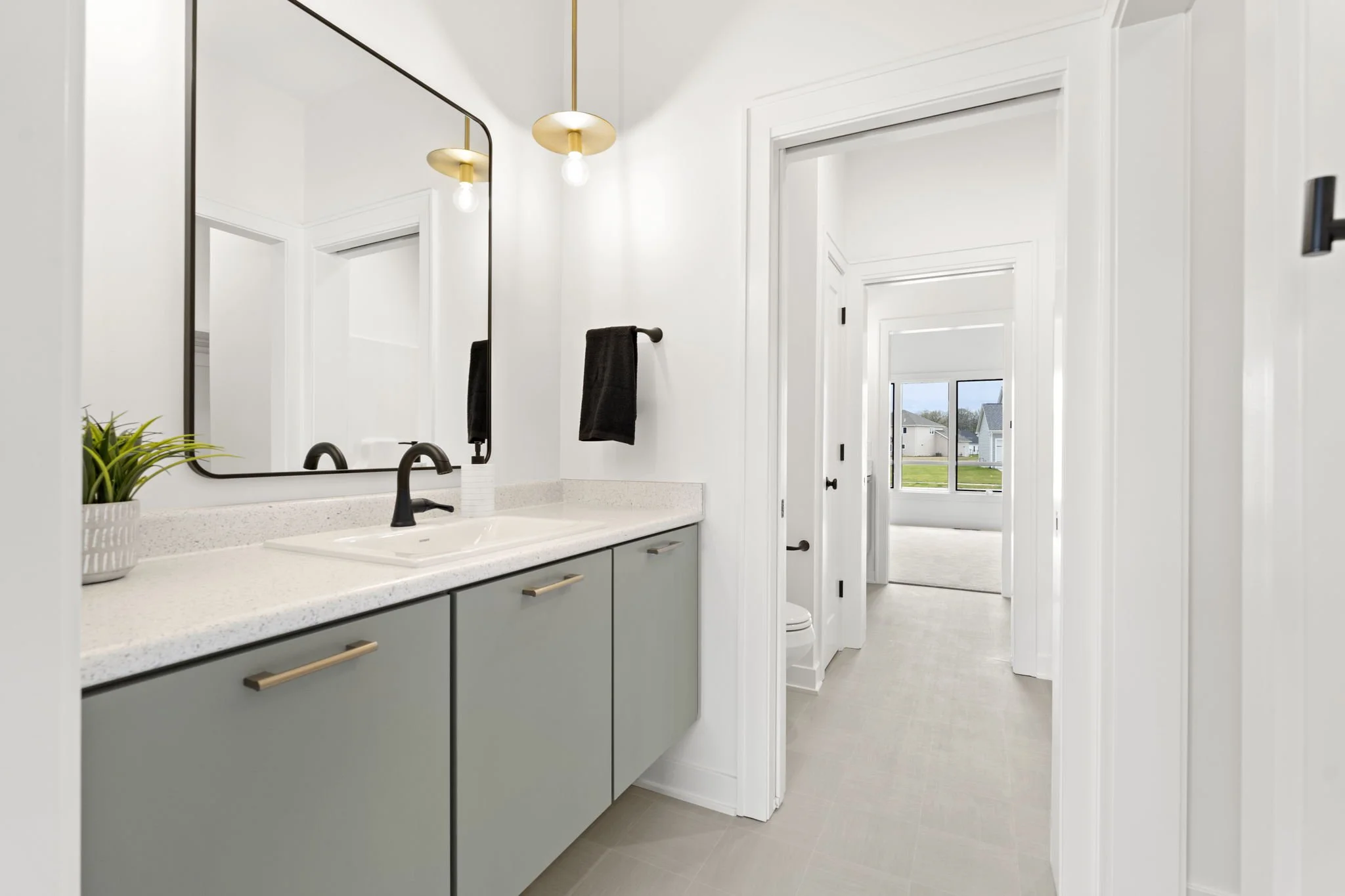 Modern bathroom with white walls, black fixtures, a large mirror, and light green cabinets. A small plant and black towel are on the countertop. Through the doorway, a visible toilet and a window showing a house and grass outside.