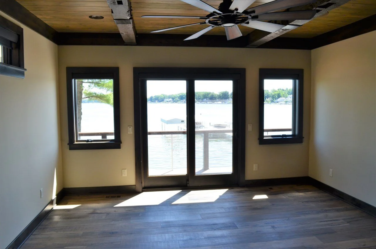 Empty room with wooden floor, cream walls, black trim, a ceiling fan, and large windows and a sliding door overlooking a lake and dock.