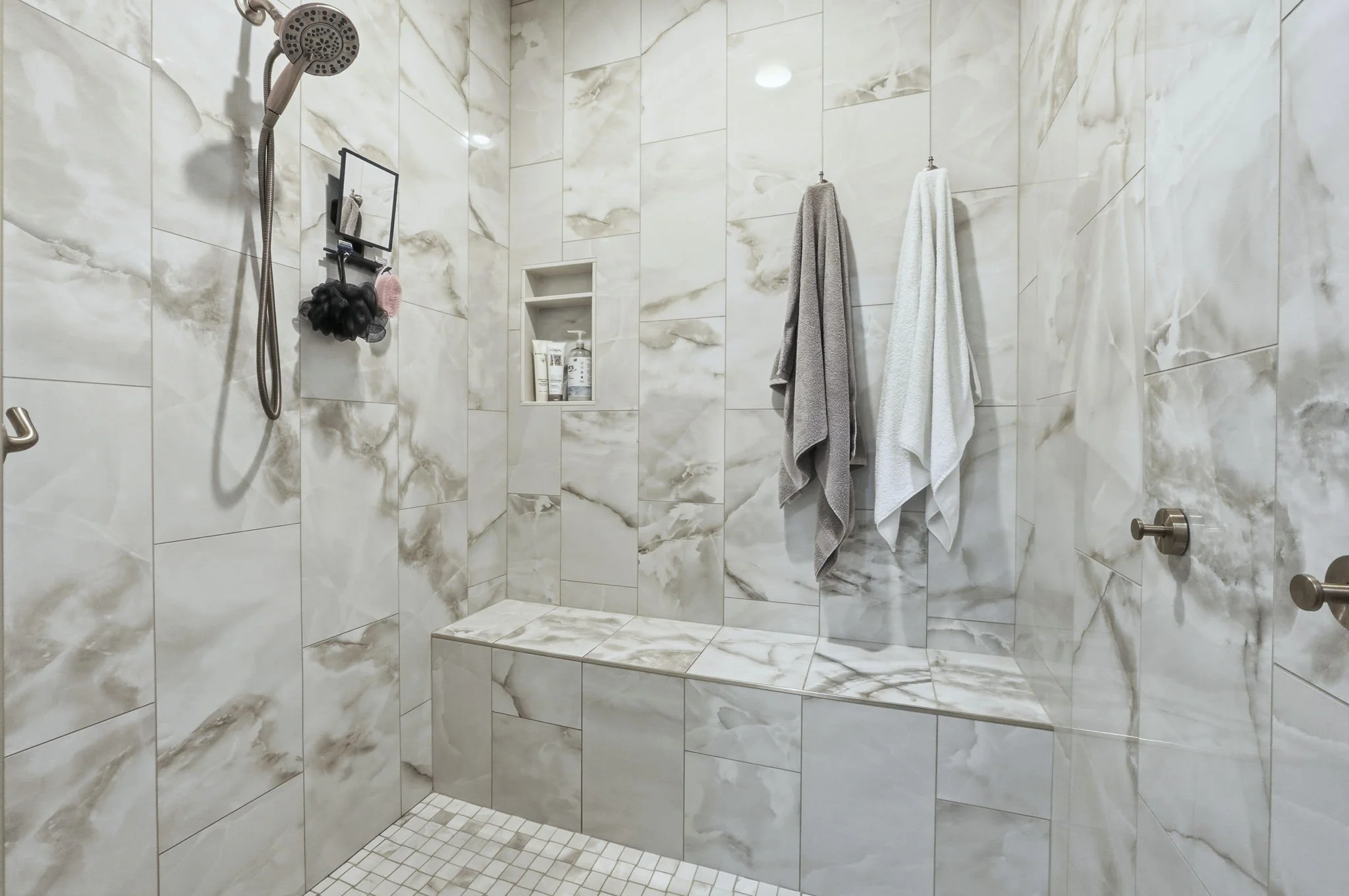 A walk-in shower with marble-look tiles on the walls and a tiled bench seat. A handheld showerhead is mounted on the wall, and there are two towels hanging on hooks. A small inset shelf holds toiletries.