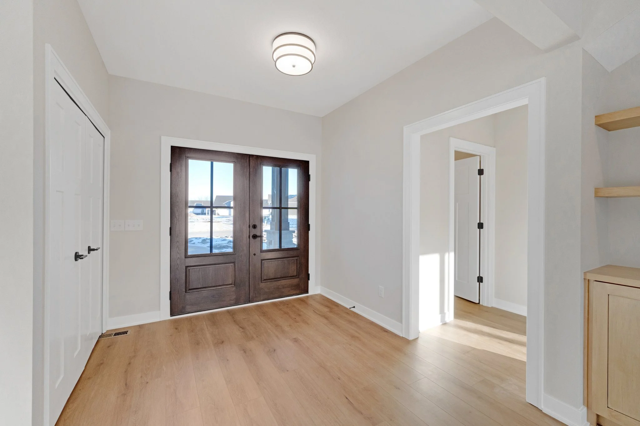 Empty room with hardwood floors, white walls, and a white ceiling with a round light fixture. Front door is dark wood with glass panels. To the left, a white closet door. To the right, an open doorway leading to another room. There are wooden shelves