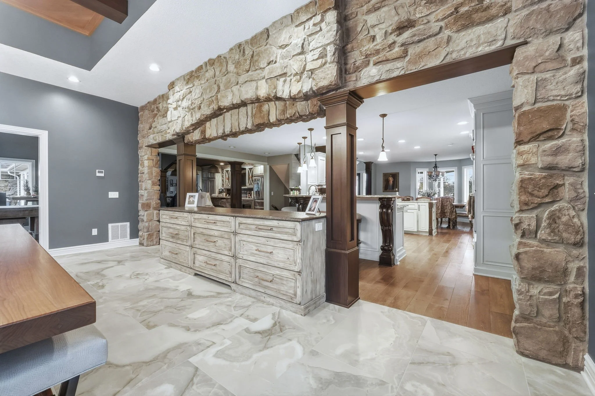 Open-concept kitchen and dining area with a stone archway, gray walls, wooden accents, and marble flooring.