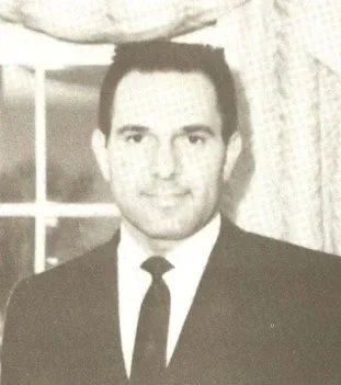 Black and white photo of a man in a suit and tie, standing indoors near a window.